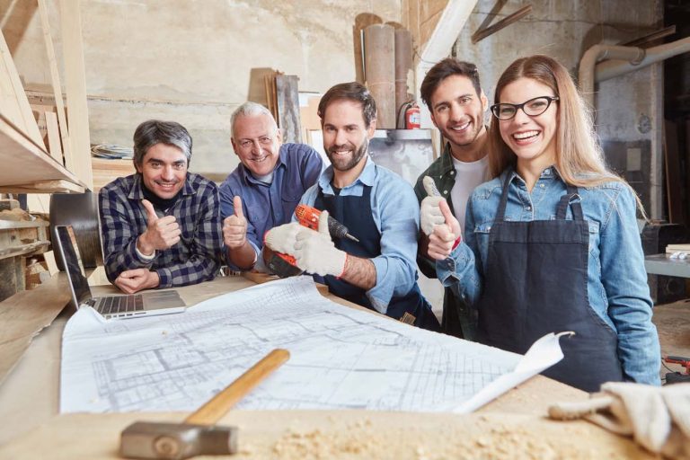 Fünf Handwerker in Arbeitskleidung zeigen Daumen hoch vor Bauplänen in einer Tischlerei.