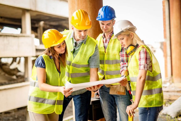 Vier Bauarbeiter mit Helmen und Warnwesten betrachten Pläne auf einer Baustelle.