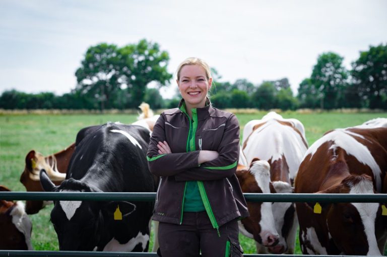 Eine Landwirtin steht lächelnd vor einer Kuhherde auf einer Wiese.